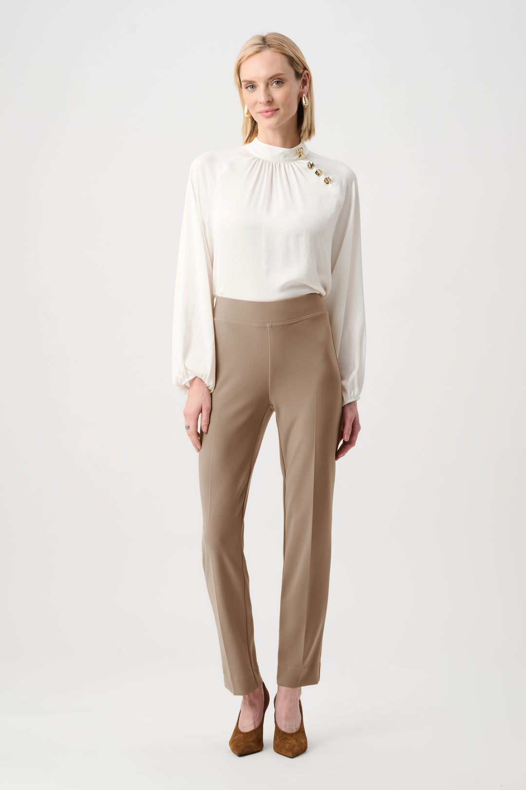 A woman modeling a white long-sleeve blouse with gold button details at the collar and waist, paired with beige straight-leg trousers and brown pointed-toe heels, against a plain light gray studio background.