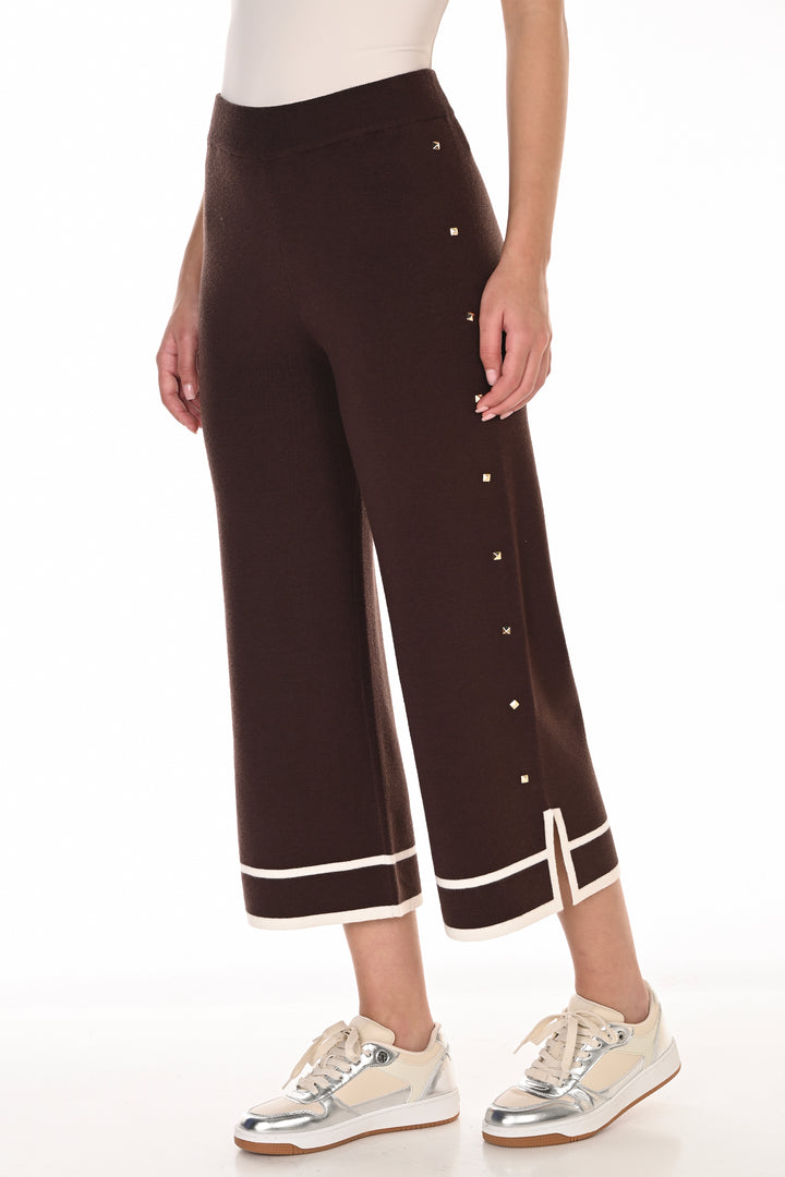 A model wearing dark brown cropped wide-leg pants with white contrast trim and silver-tone decorative buttons down the side, paired with white sneakers against a white studio background.