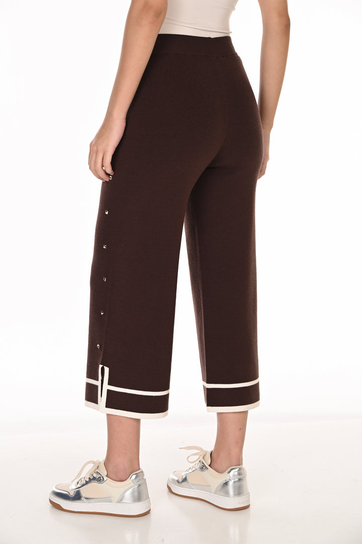 A model is shown from the back wearing dark brown cropped trousers with white contrast piping and side buttons, paired with white and silver sneakers against a white studio background.