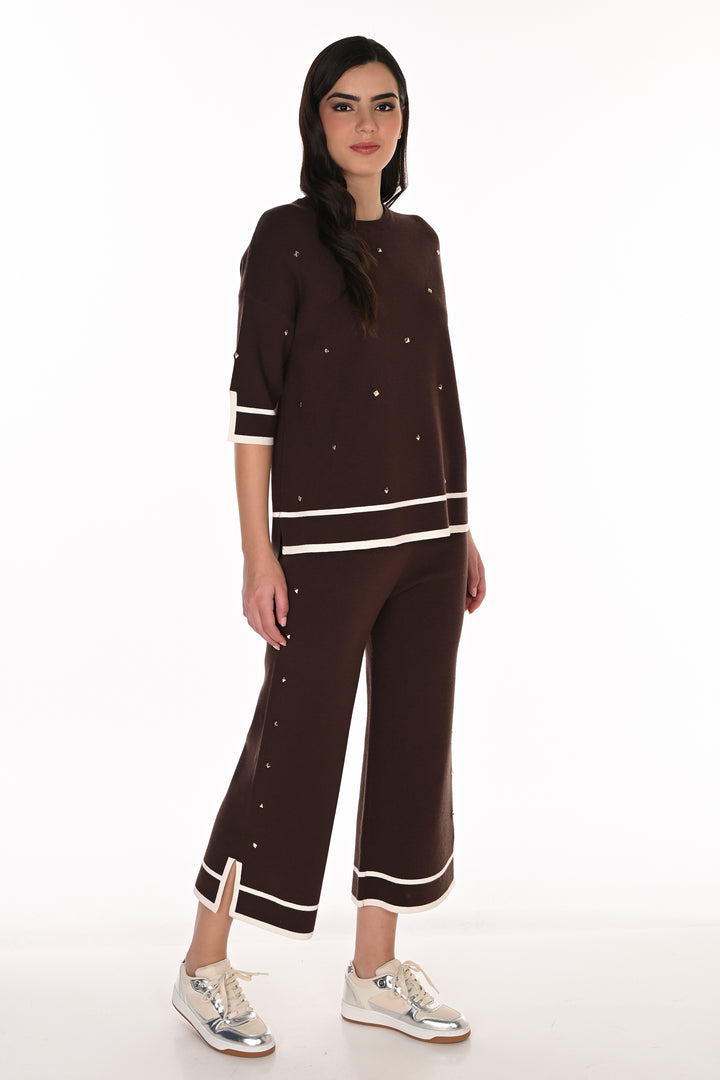 A woman modeling a dark brown knit two-piece set consisting of a short-sleeve top and matching cropped pants, both decorated with small silver rhinestones and white stripe accents, paired with silver sneakers against a white studio background.