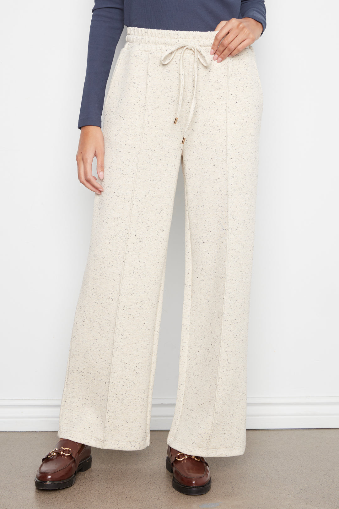 A person wearing cream-colored wide-leg drawstring pants with a subtle speckled texture, styled with a navy long-sleeve top and brown leather loafers against a white wall.