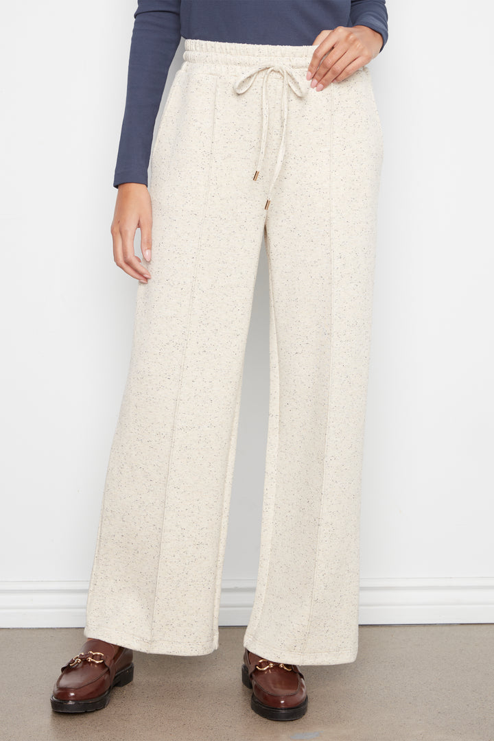 A person wearing cream-colored wide-leg drawstring pants with a subtle speckled texture, styled with a navy long-sleeve top and brown leather loafers against a white wall.