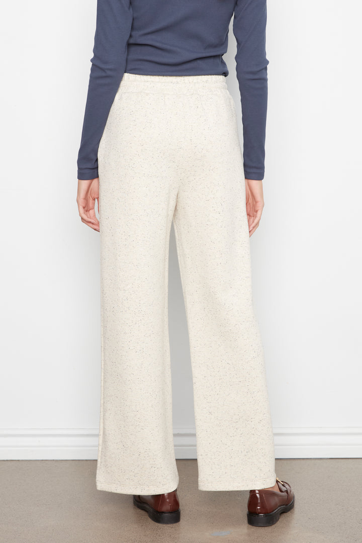 A person shown from the back wearing high-waisted, wide-leg trousers in a light cream or off-white heathered fabric, paired with a navy long-sleeve top and brown shoes, against a plain white wall.
