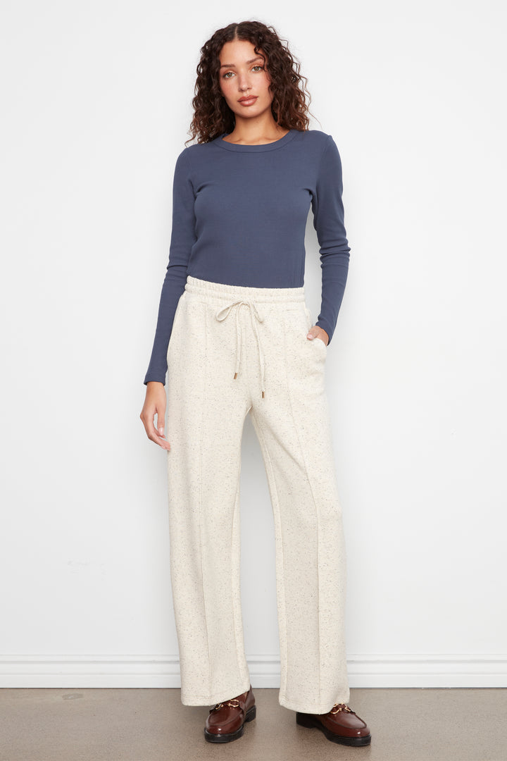 A woman modeling a slate blue long-sleeve top and cream wide-leg drawstring pants against a white wall.