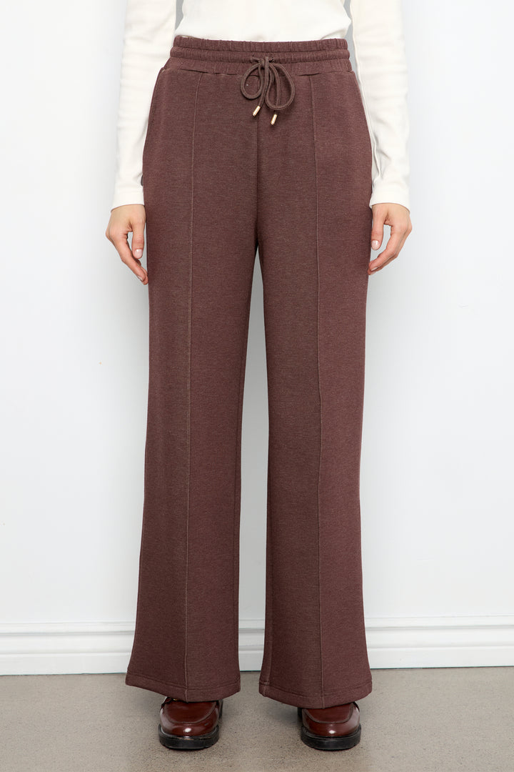 A person wearing wide-leg brown pants with a drawstring waist, paired with a white long-sleeve top and brown shoes, standing against a plain white wall.