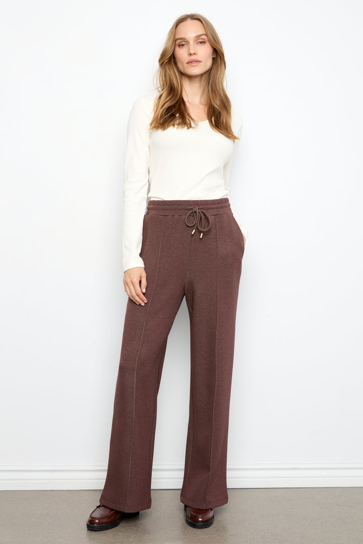 A woman modeling a pair of wide-leg brown pants with a drawstring waist, paired with a white long-sleeve top and brown shoes, standing against a plain white wall.