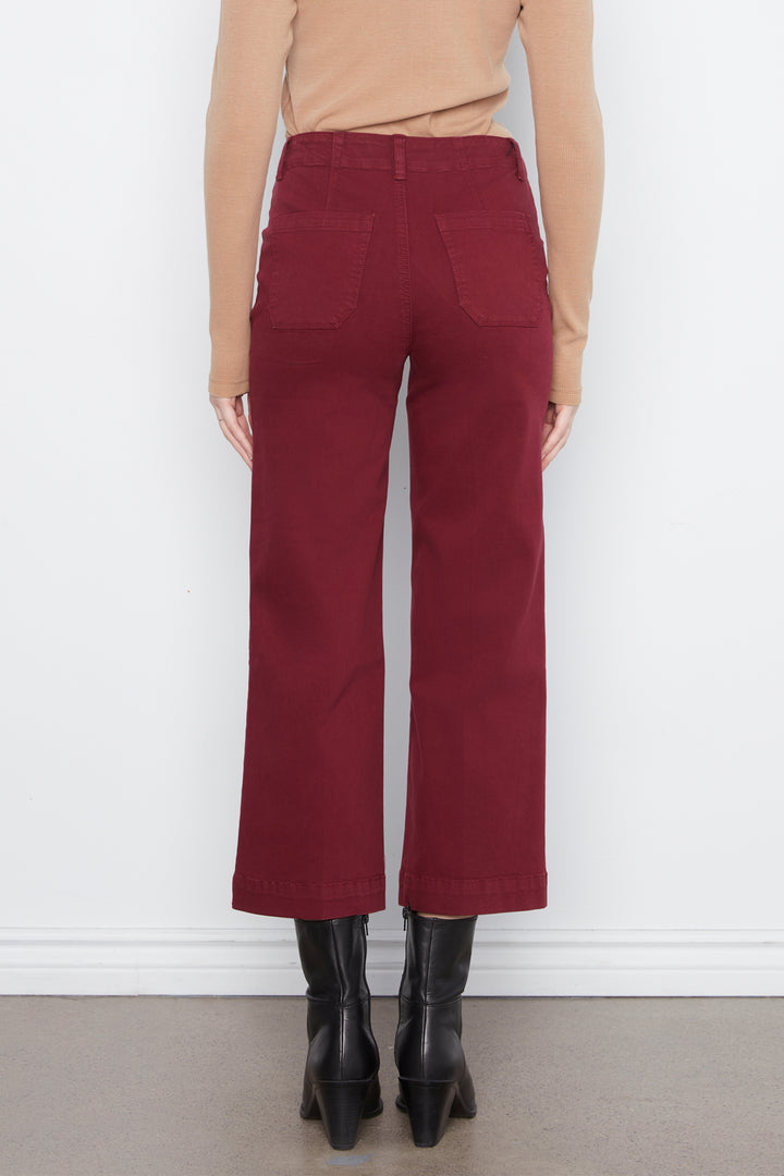 A person shown from the back wearing high-waisted, wide-leg cropped pants in a deep burgundy red, paired with a beige long-sleeve top and black ankle boots, standing against a white wall.