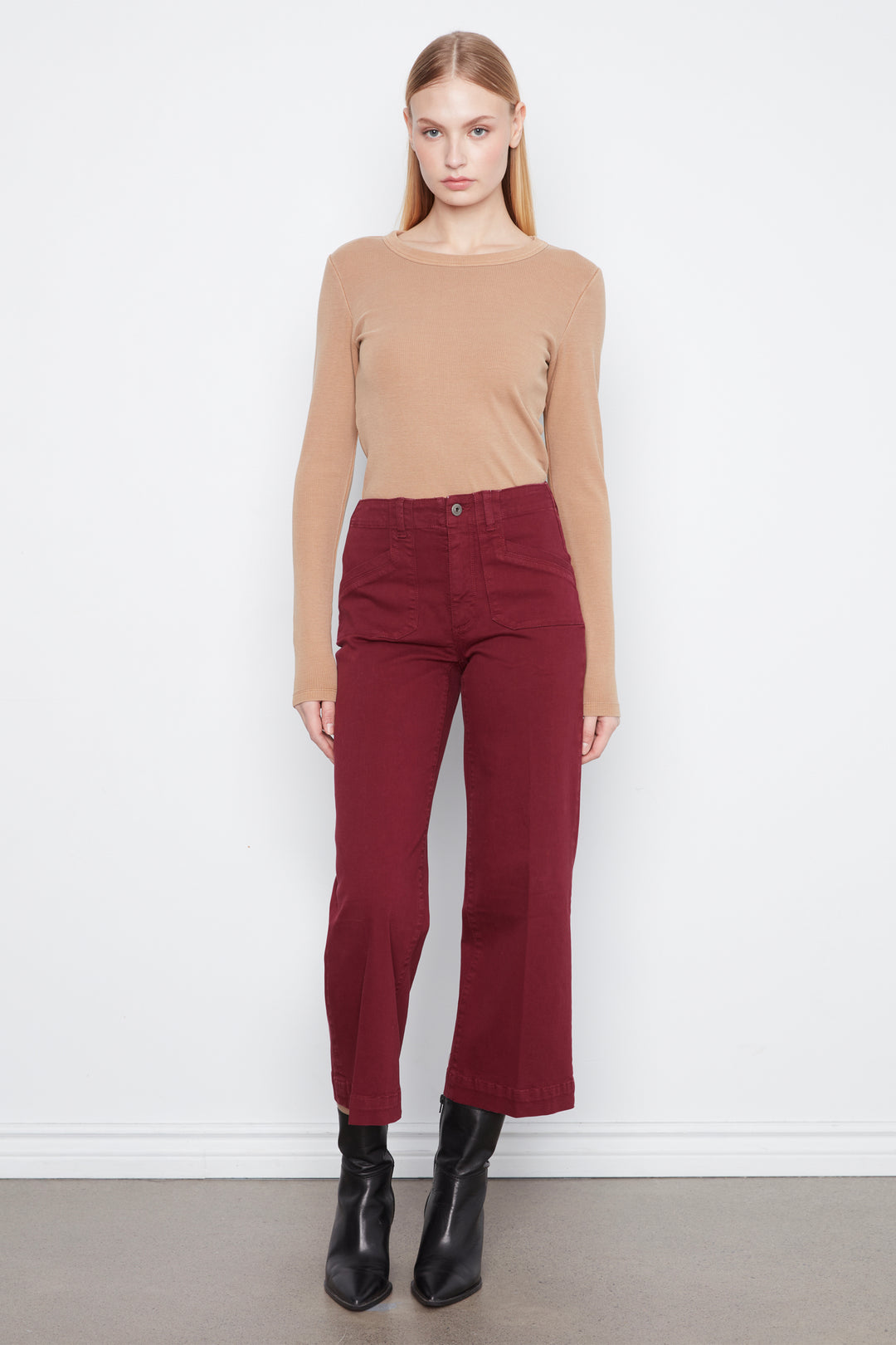 A woman stands against a white wall wearing a beige long-sleeve knit top and dark red cropped pants with black ankle boots.