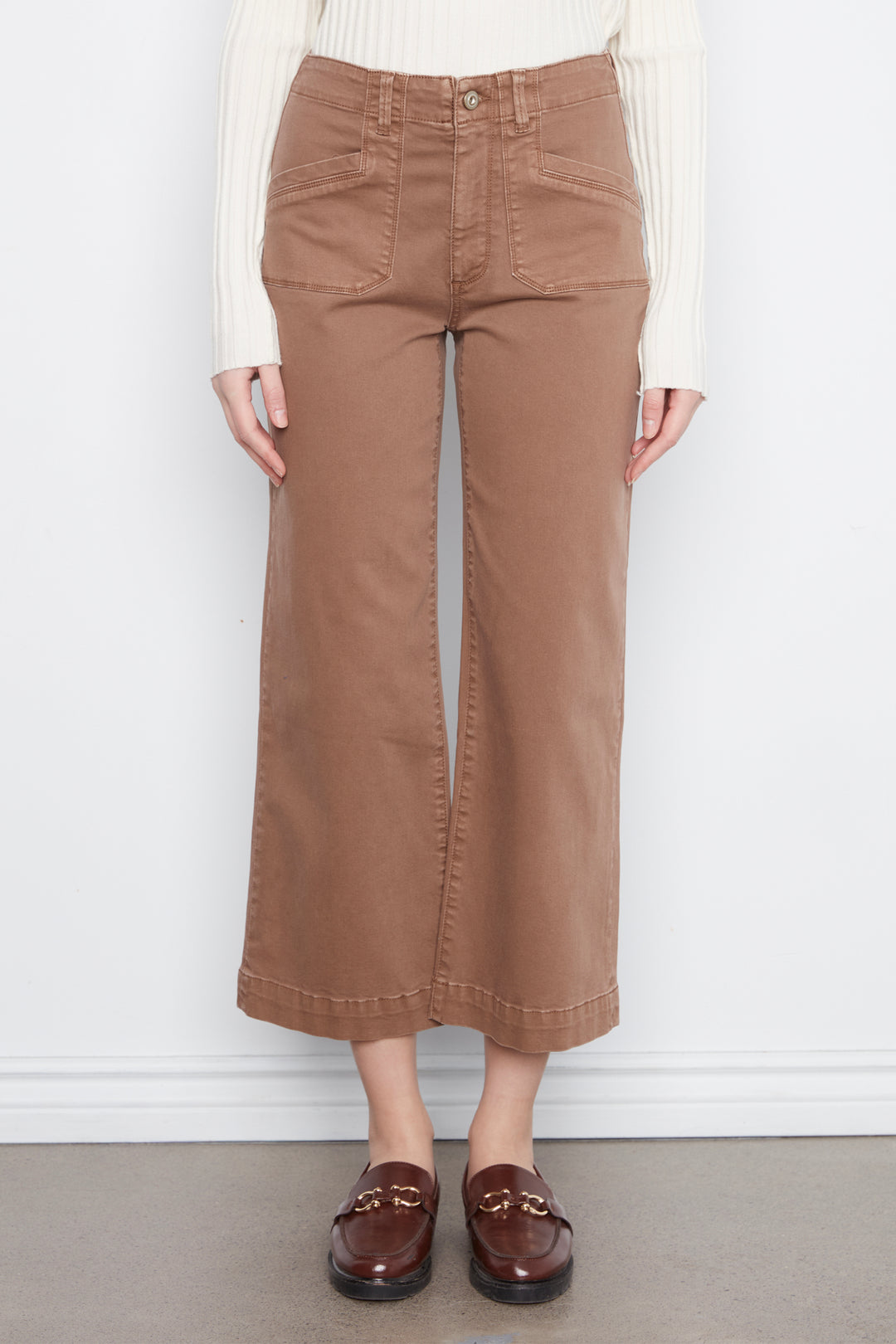 A person wearing brown wide-leg cropped pants with a cream ribbed long-sleeve top and dark brown loafers, standing against a white wall.