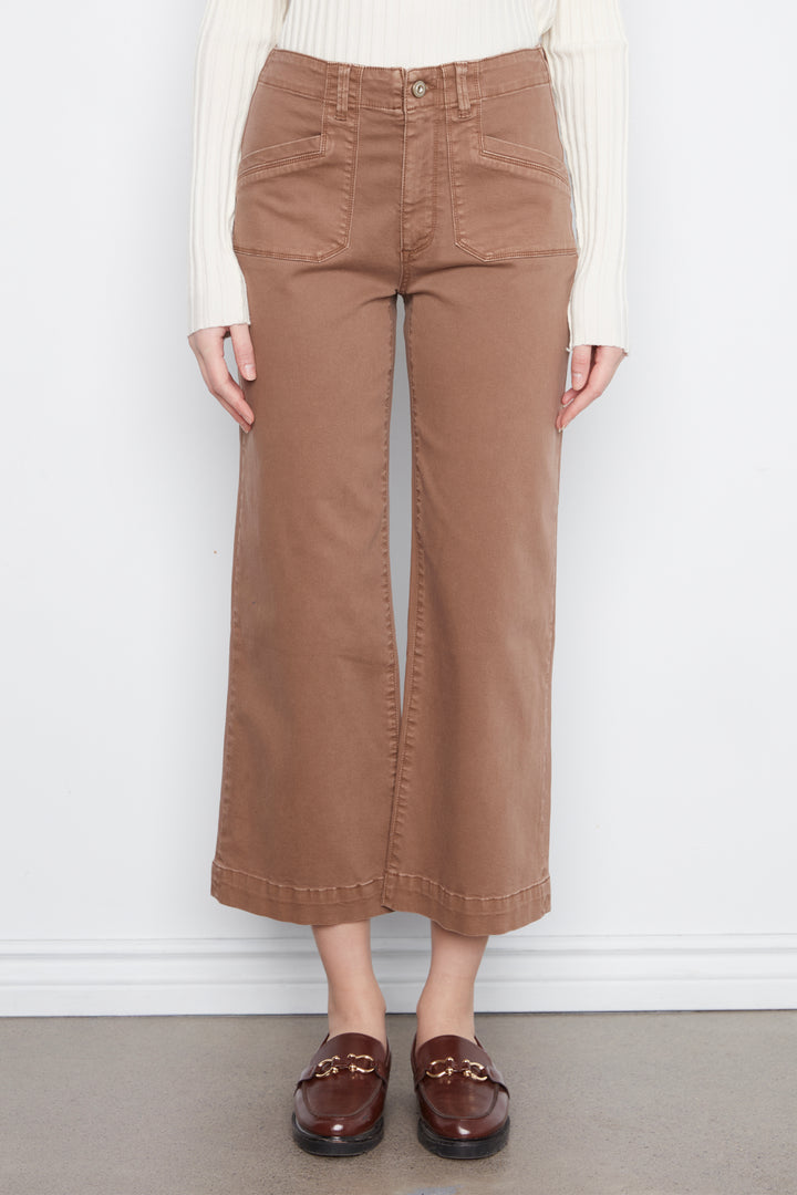 A person wearing brown wide-leg cropped pants with a cream ribbed long-sleeve top and dark brown loafers, standing against a white wall.