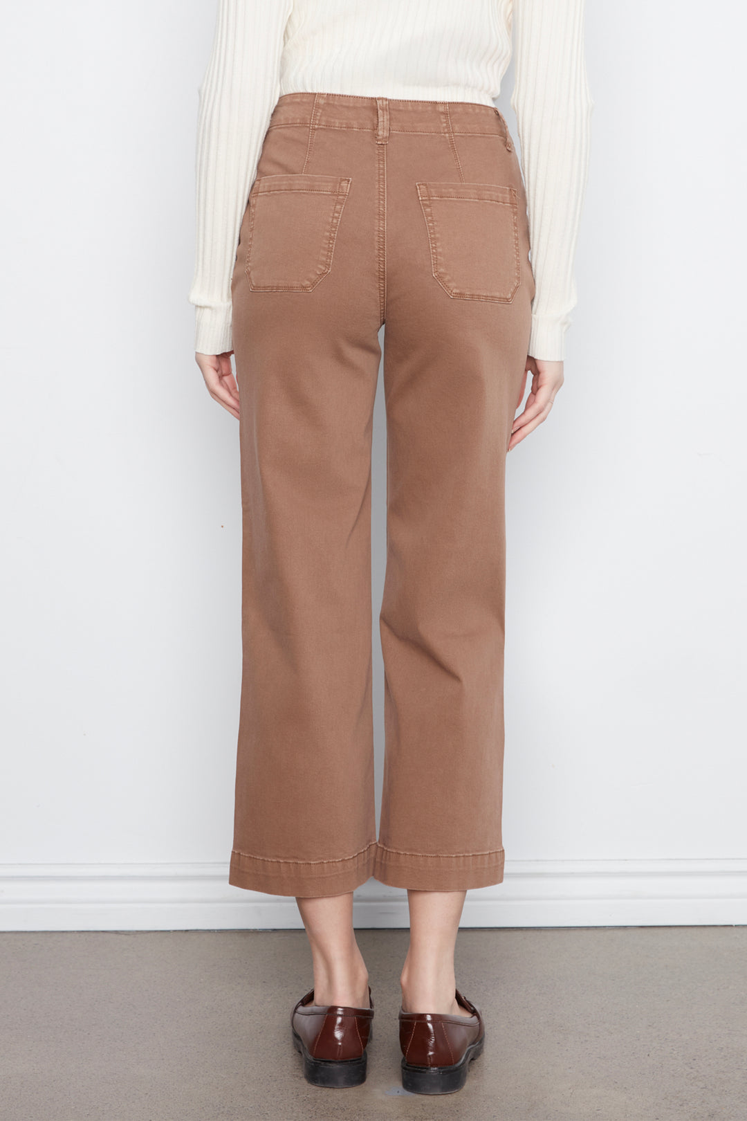 A person shown from the back wearing tan cropped wide-leg pants with back patch pockets, a cream ribbed sweater, and dark brown loafers, standing against a plain white wall.
