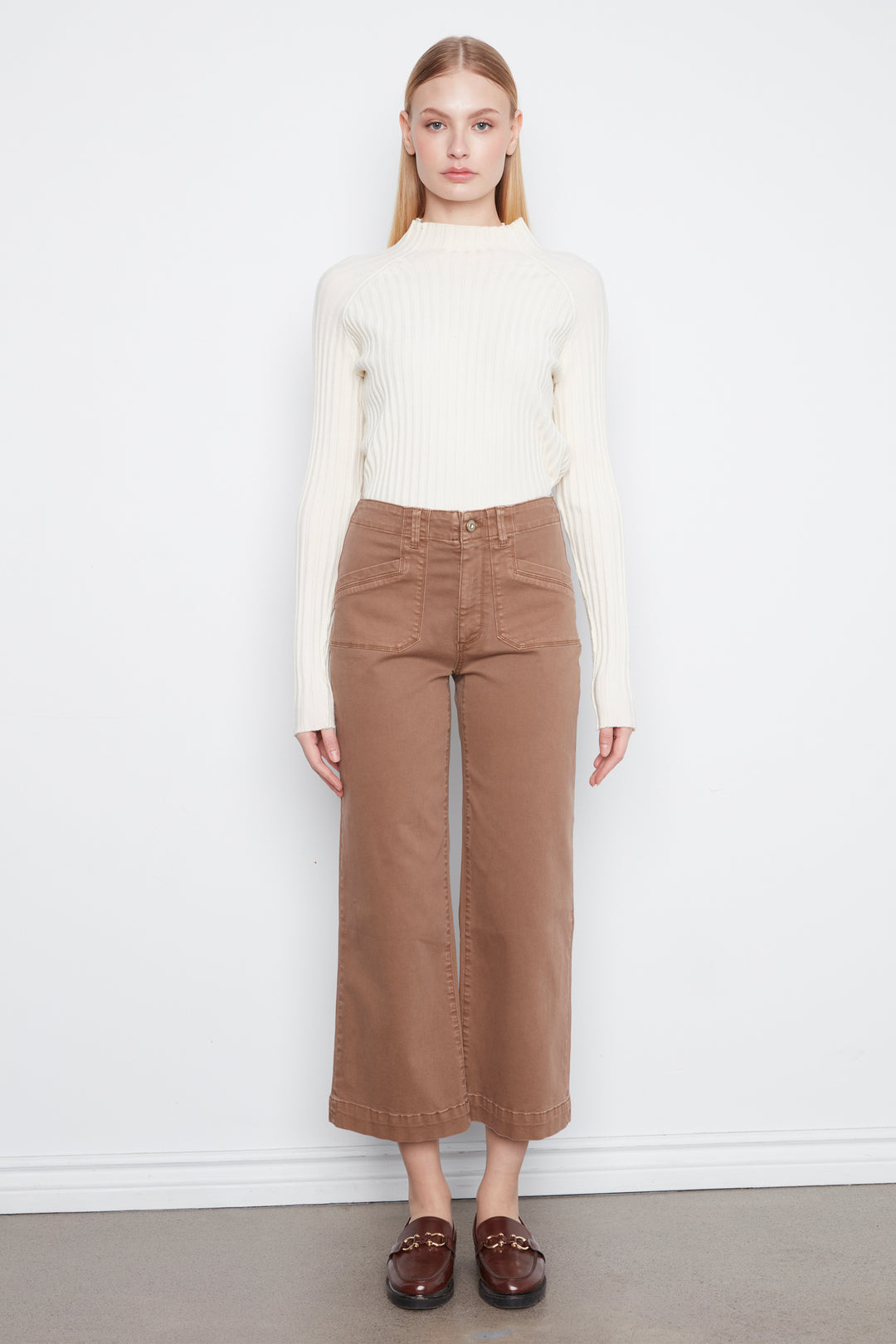 A model stands against a white wall wearing a cream ribbed mock-neck long-sleeve sweater, brown cropped wide-leg jeans, and dark brown leather loafers.
