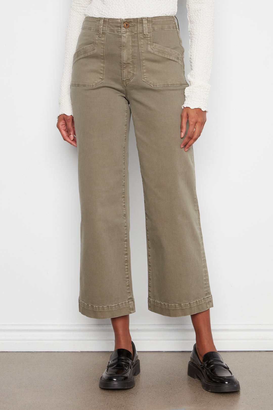 A model wearing a pair of olive green cropped wide-leg jeans with a white long-sleeve top and black leather loafers, standing against a white wall.