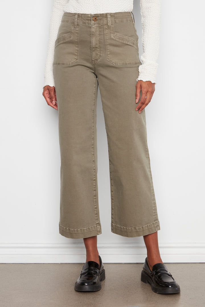 A model wearing a pair of olive green cropped wide-leg jeans with a white long-sleeve top and black leather loafers, standing against a white wall.