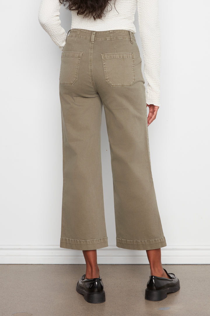 A person shown from the back wearing high-waisted, wide-leg cropped pants in a light khaki or taupe color, paired with a white ribbed long-sleeve top and black loafers, against a plain white wall.