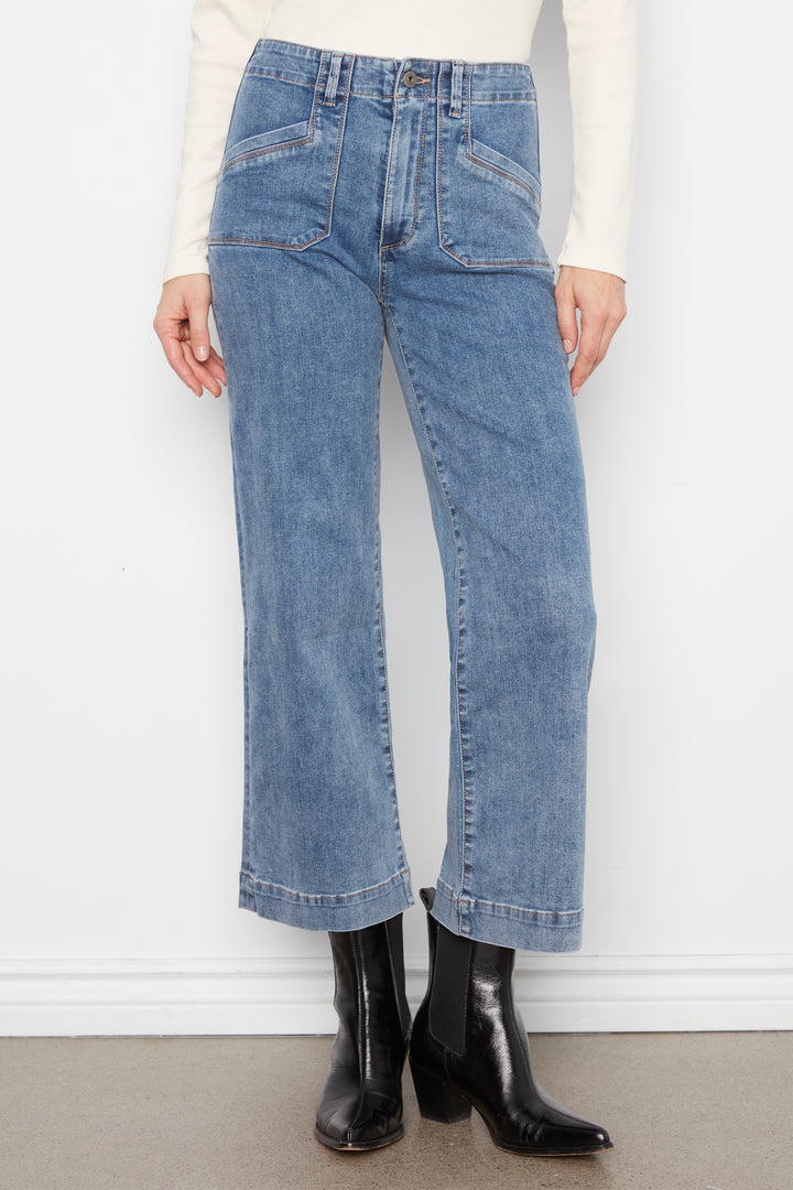 A model wearing light blue wide-leg cropped jeans, paired with a cream long-sleeve top and black leather ankle boots, standing against a white wall.