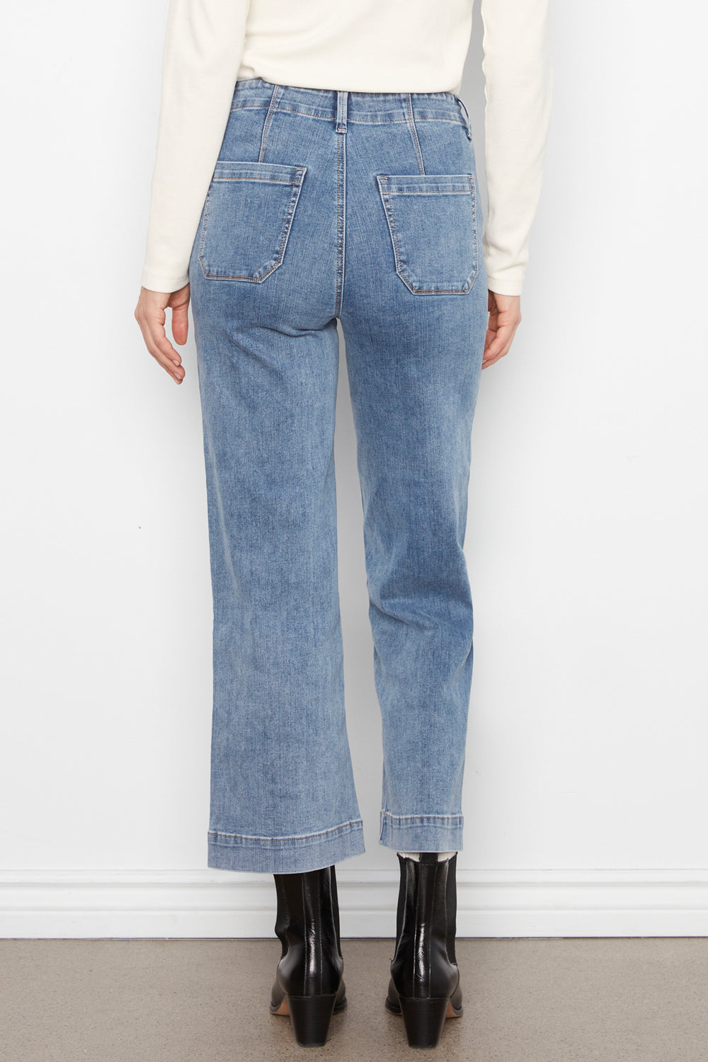 A person shown from the back wearing light-wash blue cropped jeans with a straight-to-slightly-flared leg, paired with a cream top and black ankle boots against a plain white wall.