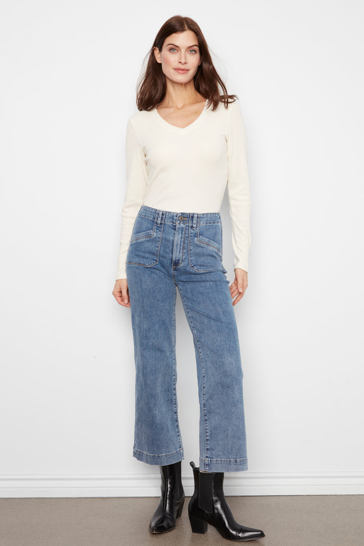 A woman modeling a cream long-sleeve V-neck top and high-waisted, wide-leg blue denim jeans with black ankle boots, standing against a plain white wall.