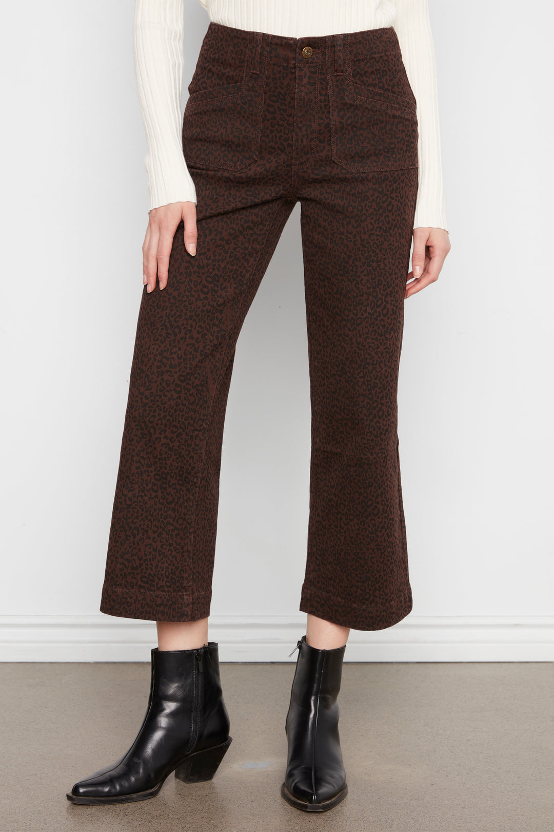 A model wearing dark brown cropped pants with a subtle leopard print, styled with a cream ribbed sweater and black leather ankle boots against a white wall.