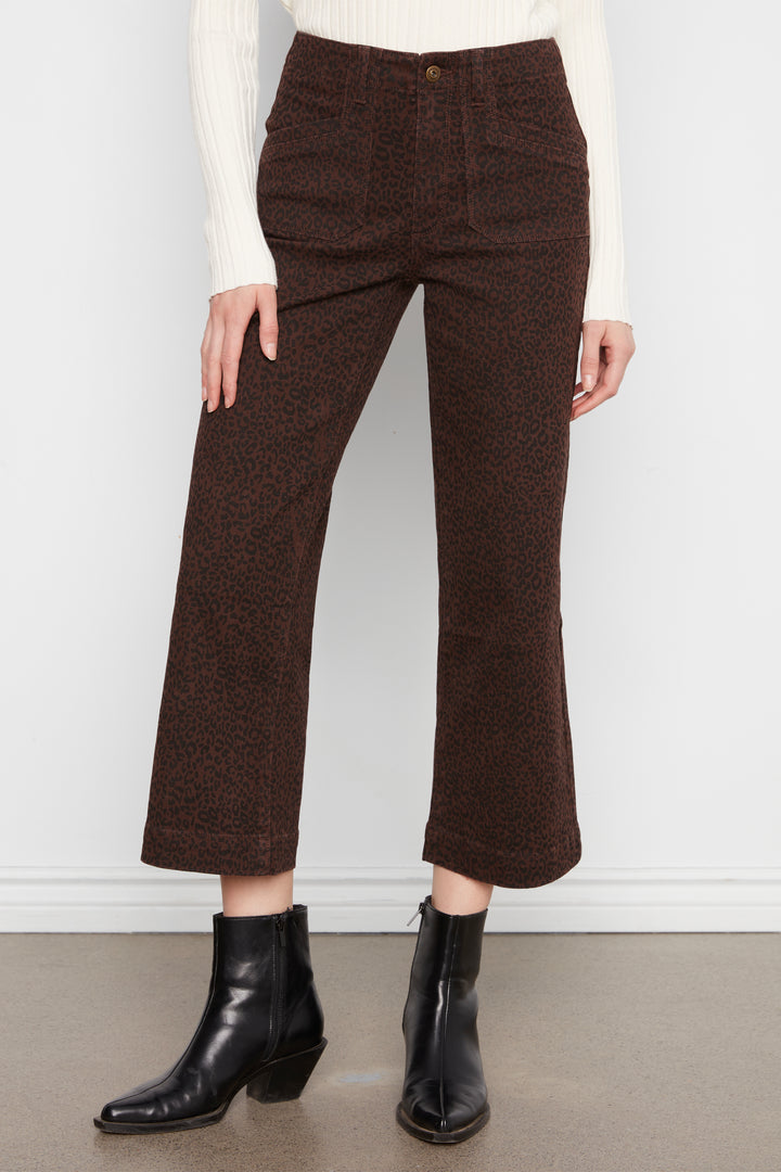 A model wearing dark brown cropped pants with a subtle leopard print, styled with a cream ribbed sweater and black leather ankle boots against a white wall.
