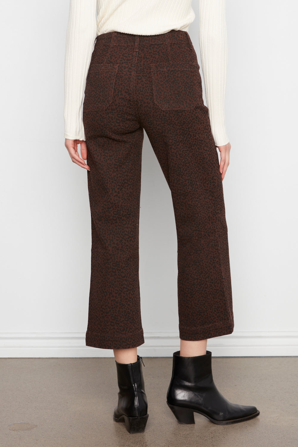 A person shown from the back wearing dark brown textured cropped trousers, a cream ribbed sweater, and black ankle boots against a plain light gray wall.