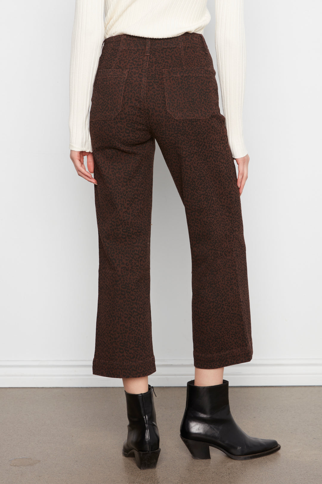 A person shown from the back wearing dark brown textured cropped trousers, a cream ribbed sweater, and black ankle boots against a plain light gray wall.