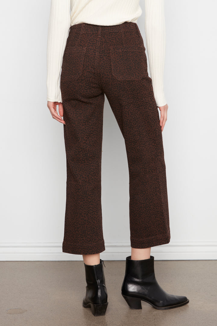 A person shown from the back wearing dark brown textured cropped trousers, a cream ribbed sweater, and black ankle boots against a plain light gray wall.