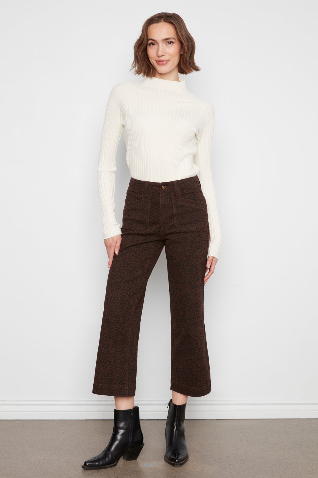 A woman modeling a cream ribbed mock-neck sweater and dark brown cropped wide-leg trousers with black ankle boots, standing against a white wall.