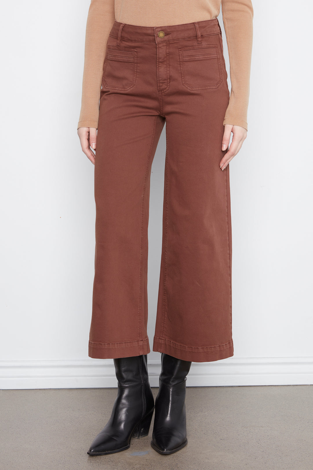 A model wearing a pair of rust-brown wide-leg cropped pants, styled with a tan long-sleeve top and black leather ankle boots, standing against a white wall.