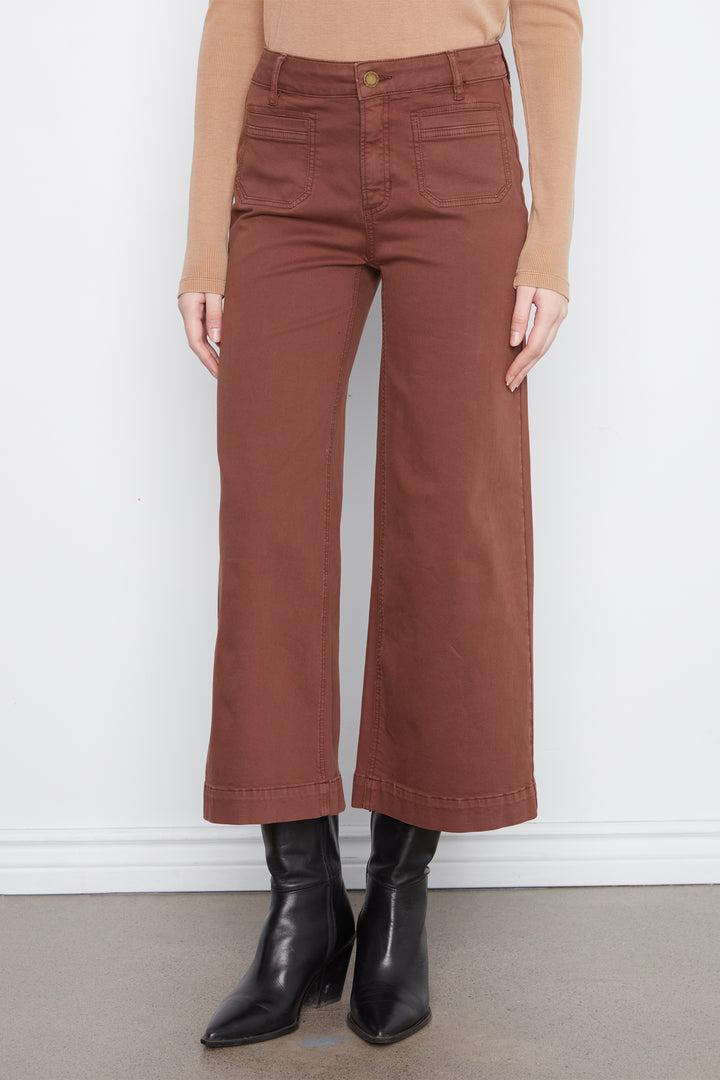 A model wearing a pair of rust-brown wide-leg cropped pants, styled with a tan long-sleeve top and black leather ankle boots, standing against a white wall.