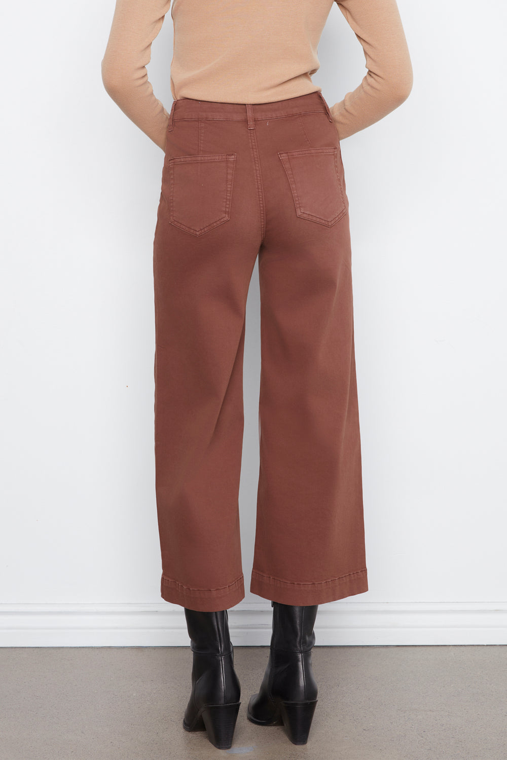 A person shown from the back wearing rust-brown wide-leg cropped pants, a beige long-sleeve top, and black ankle boots against a plain white wall.