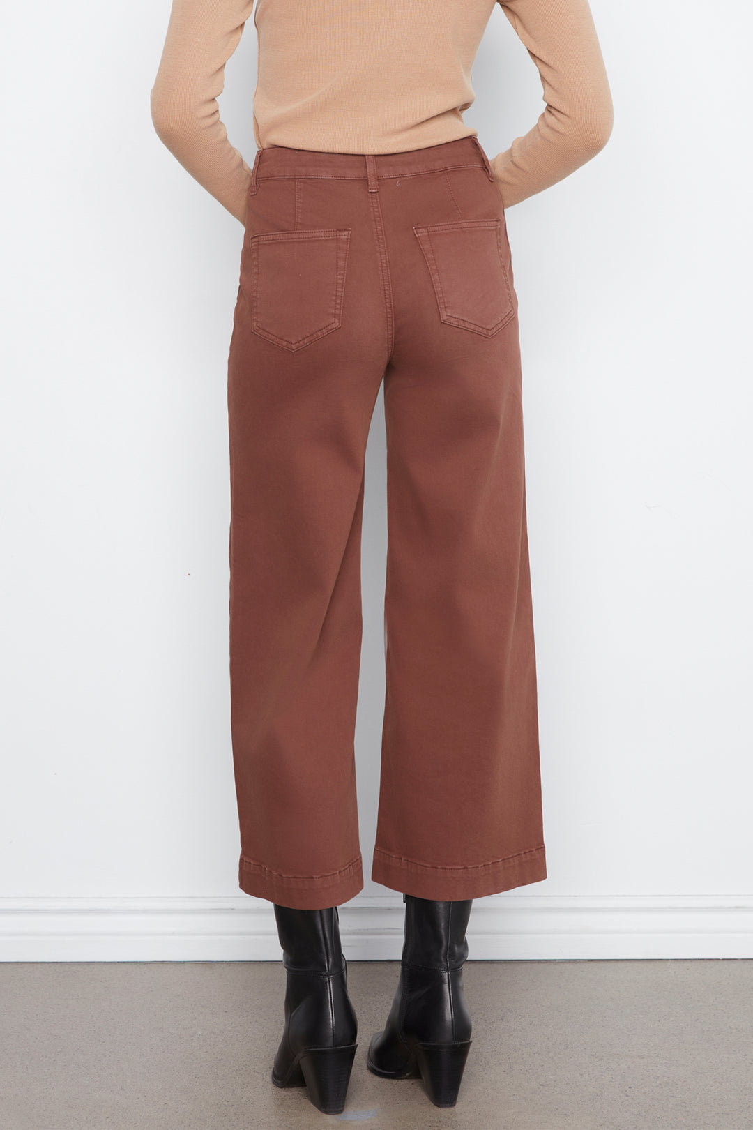 A person shown from the back wearing rust-brown wide-leg cropped pants, a beige long-sleeve top, and black ankle boots against a plain white wall.
