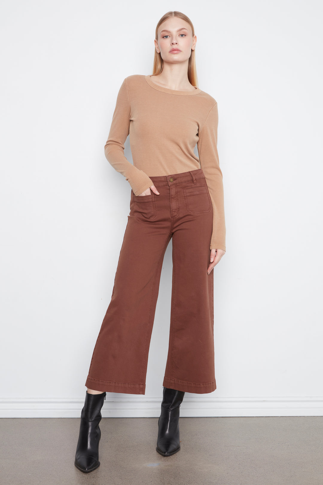 A woman modeling a tan long-sleeve knit top and rust-brown wide-leg cropped pants with black ankle boots, standing against a plain white wall.