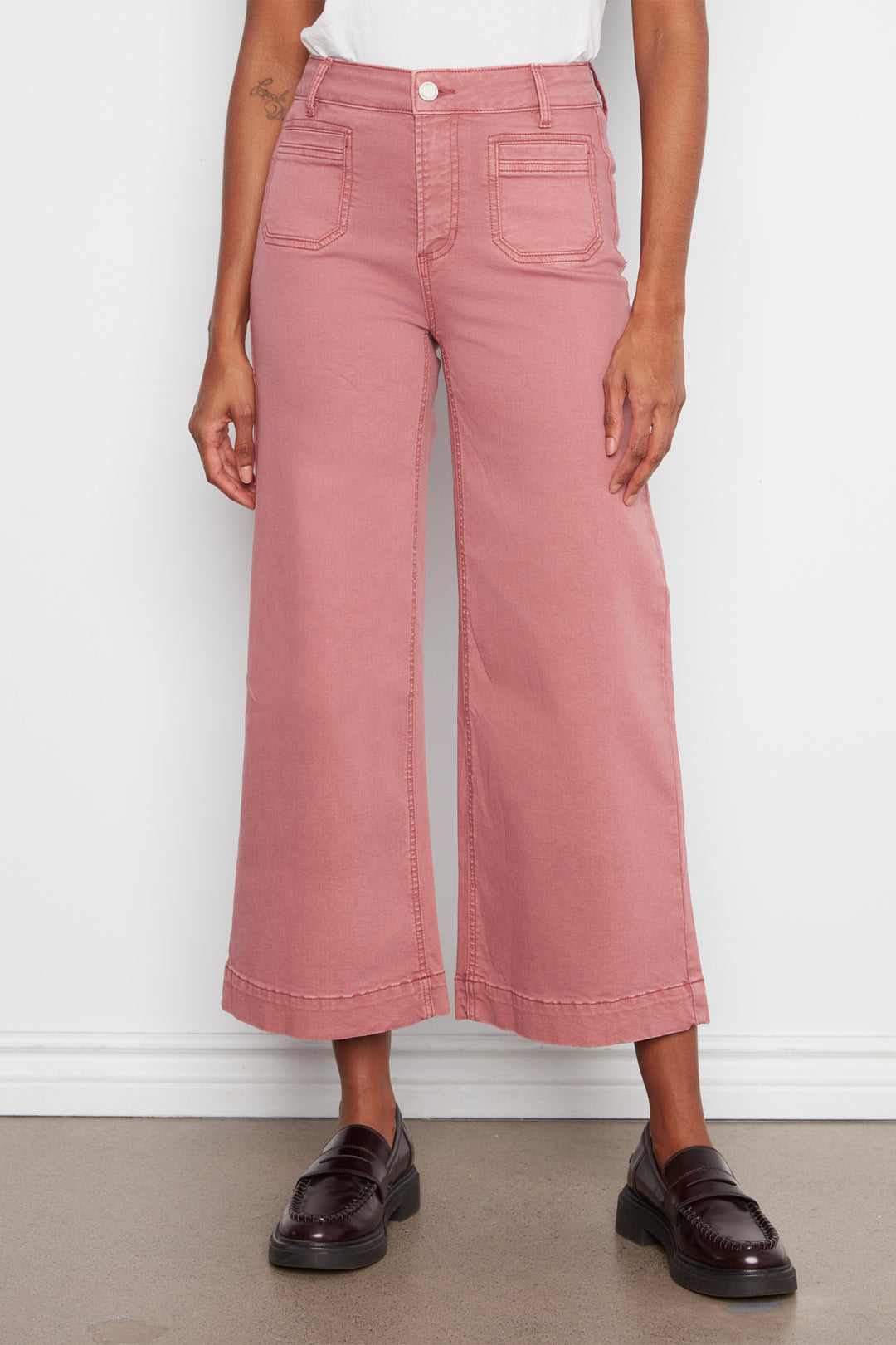 A model wearing dusty rose wide-leg cropped jeans with a white top and dark brown leather loafers, standing against a white wall.