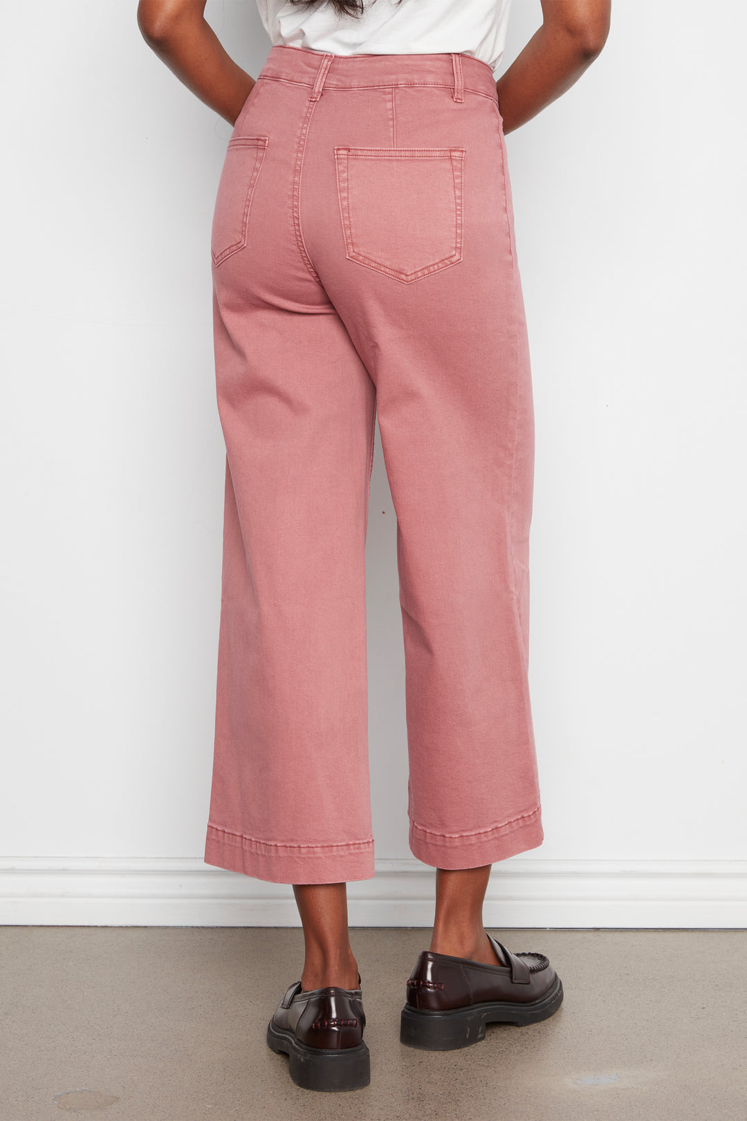 A person shown from the back wearing dusty rose cropped wide-leg jeans with a white top and dark brown leather loafers, standing against a white wall.