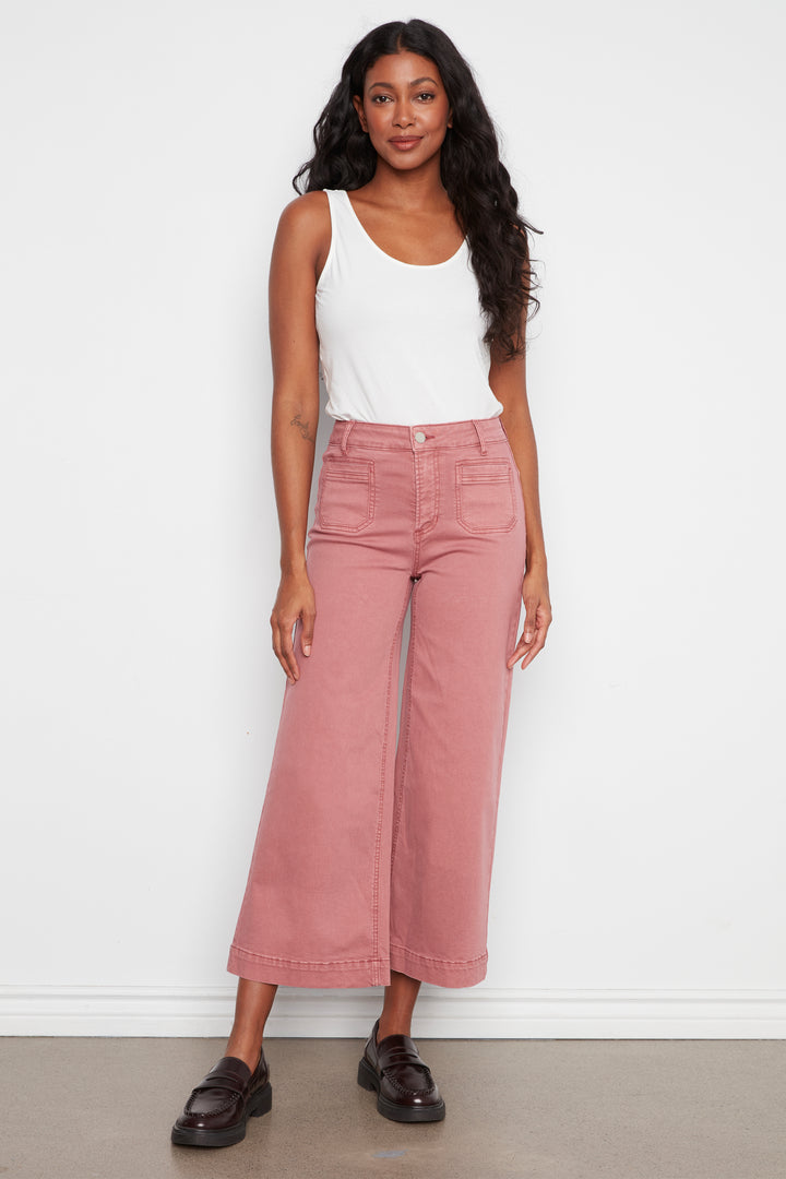 A woman modeling dusty rose wide-leg cropped jeans with a white tank top and dark brown loafers, standing against a plain white wall.