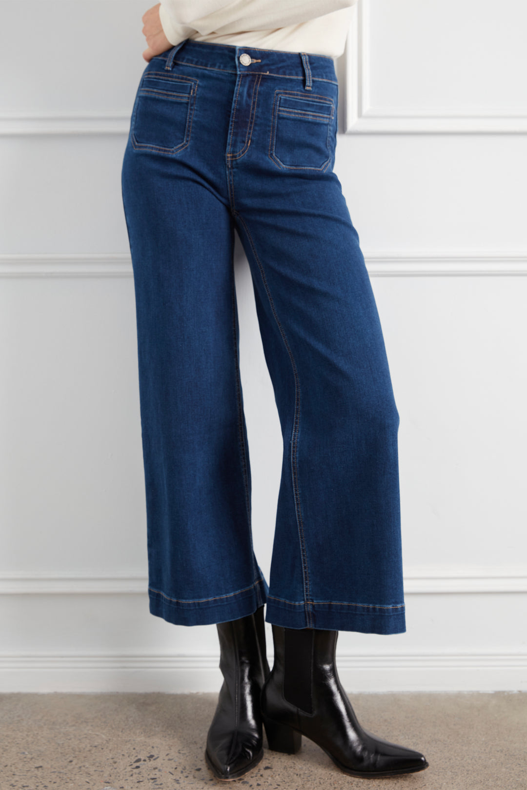 A person wearing dark blue wide-leg cropped jeans with a cream top and black ankle boots, standing against a white paneled wall.