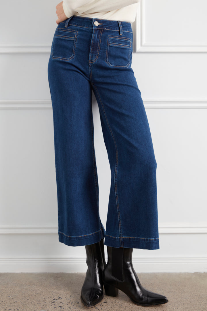 A person wearing dark blue wide-leg cropped jeans with a cream top and black ankle boots, standing against a white paneled wall.
