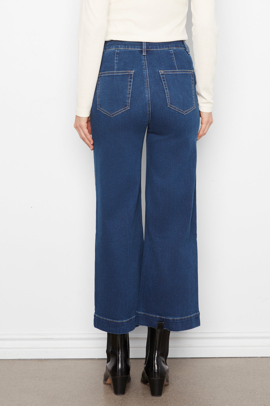 A person shown from the back wearing dark blue wide-leg cropped jeans with contrast stitching, a cream long-sleeve top, and black ankle boots against a white wall.