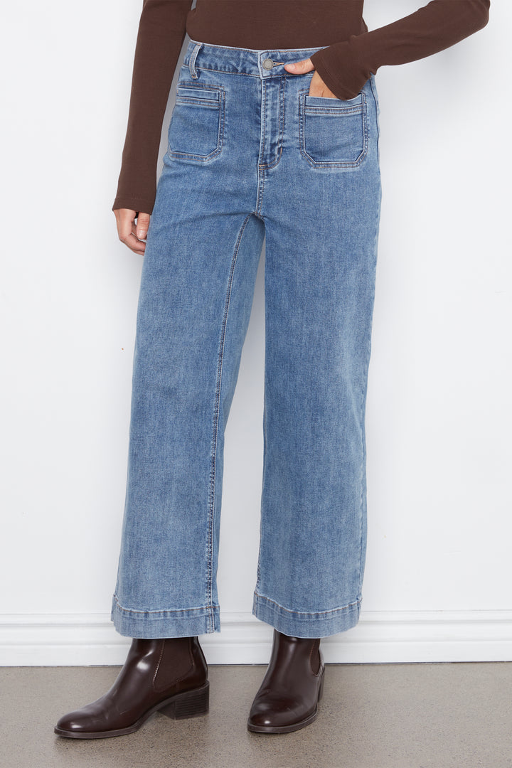 A person wearing light blue wide-leg denim jeans with a brown long-sleeve top and dark brown ankle boots, standing against a white wall.