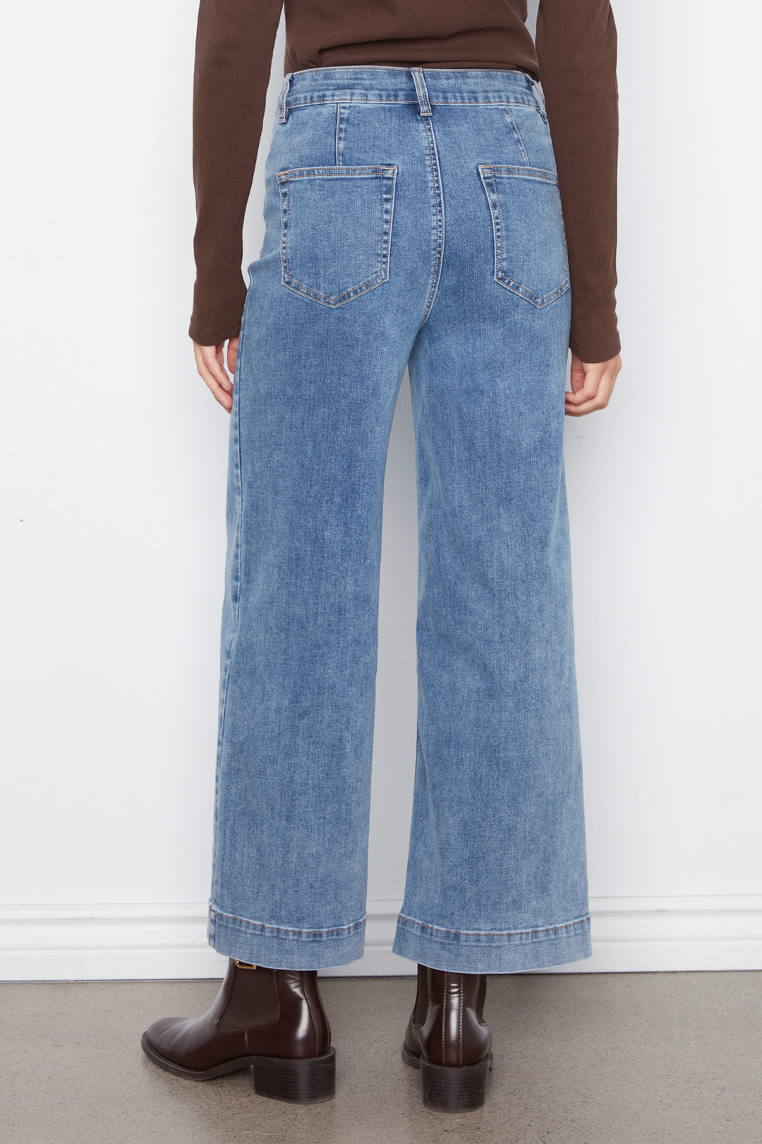A person shown from the back wearing light-wash wide-leg denim jeans, a brown long-sleeve top, and dark brown ankle boots against a plain white wall.