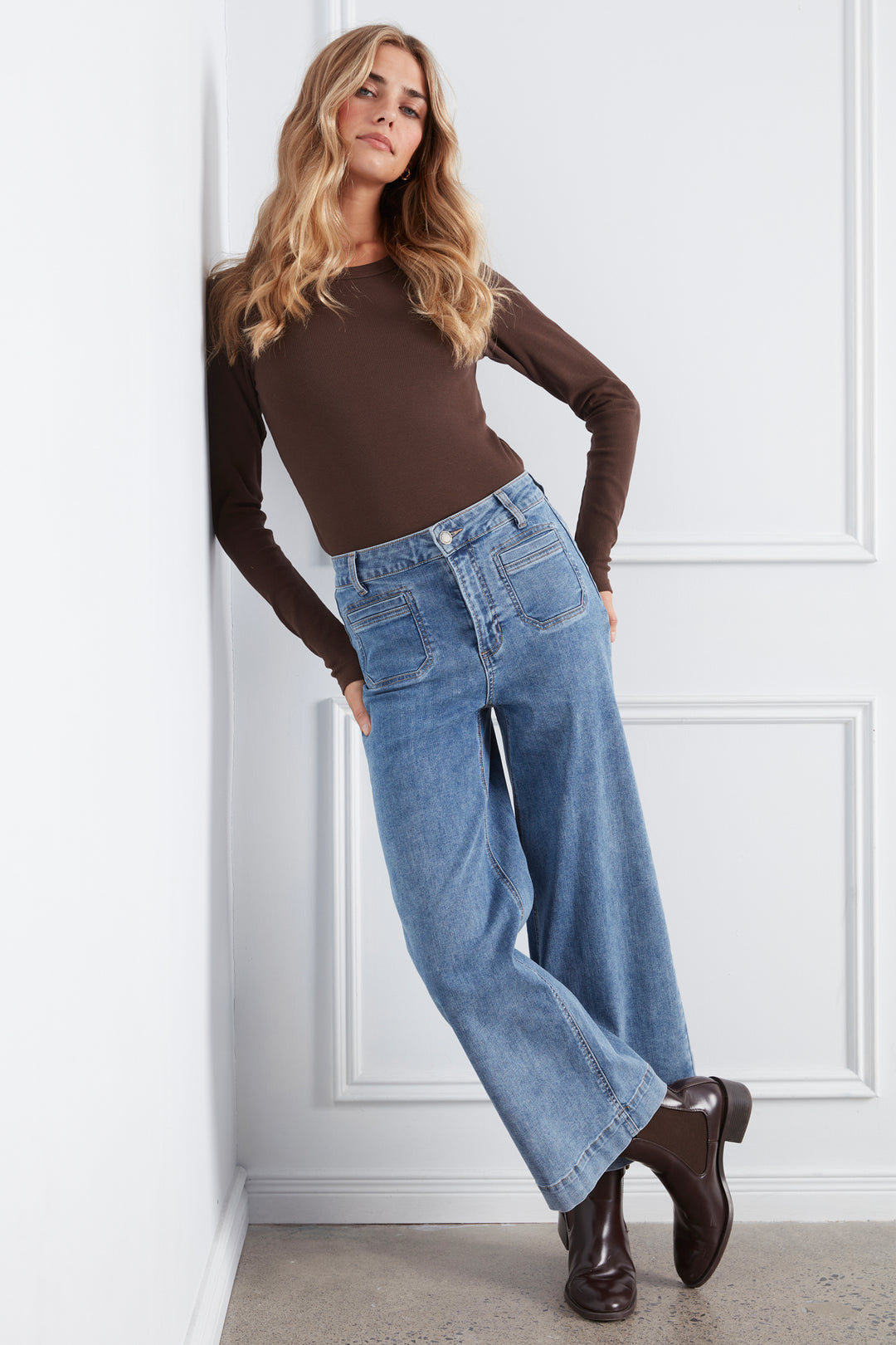 A woman modeling a brown long-sleeve top and high-waisted, wide-leg light blue jeans, standing in a white paneled room.
