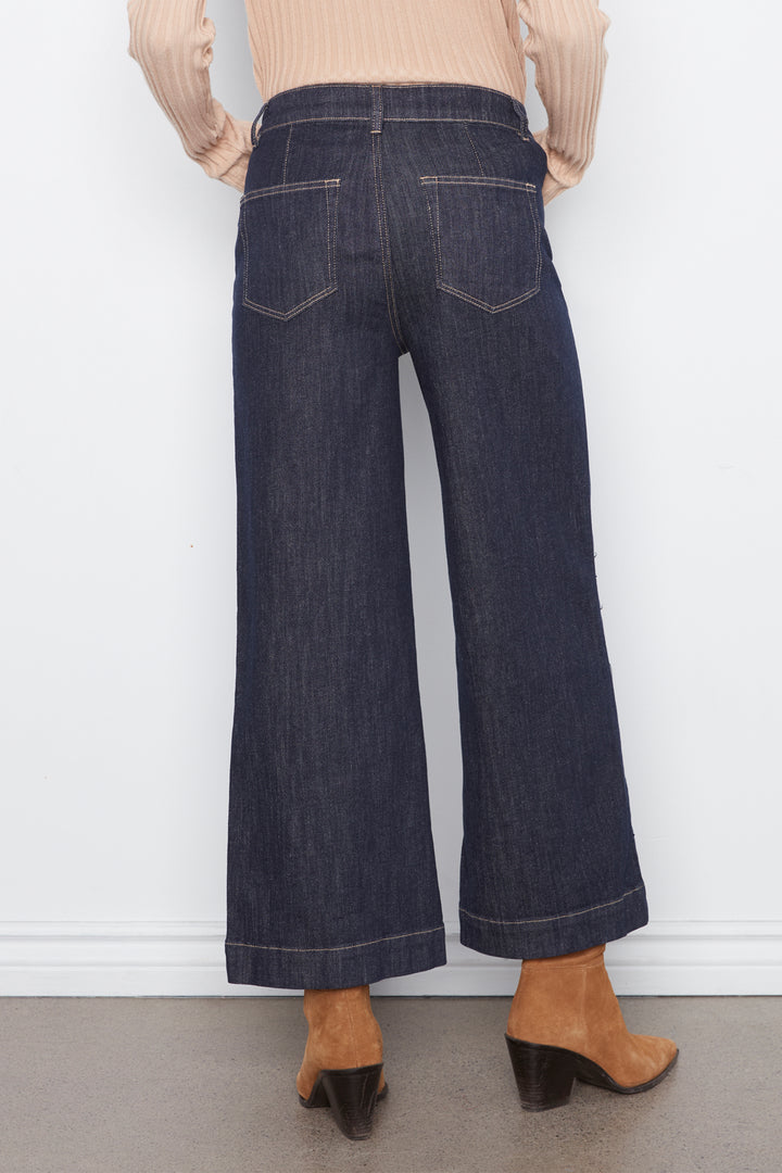 Back view of a person wearing dark blue wide-leg cropped jeans with a beige ribbed top and tan heeled mule sandals, standing against a white wall.