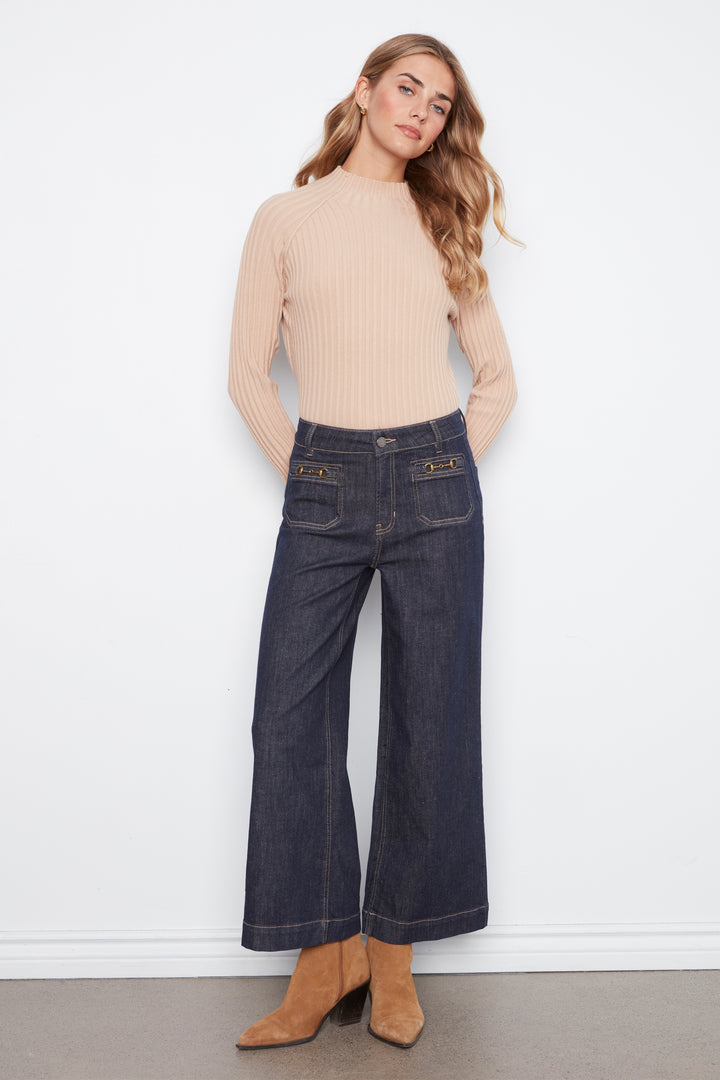 A woman stands against a white wall wearing a beige ribbed mock-neck long-sleeve top, dark blue wide-leg denim jeans with gold button accents, and tan ankle boots.