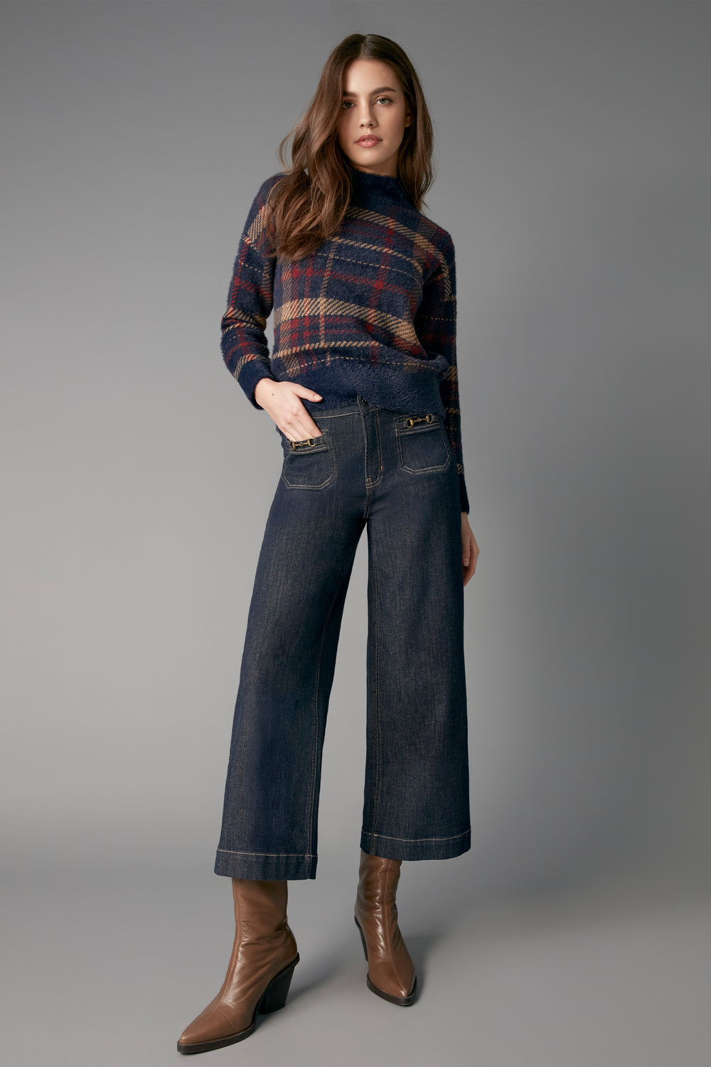 A woman modeling a dark blue plaid turtleneck sweater, dark wide-leg cropped jeans, and brown leather wedge boots against a solid gray studio background.
