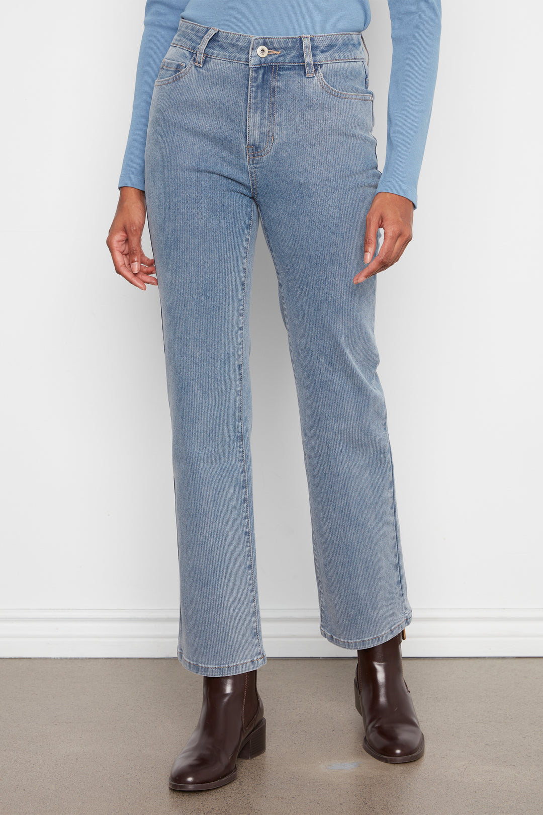 A person wearing light-wash blue straight-leg jeans with a light blue long-sleeve top and dark brown ankle boots, standing against a white wall.