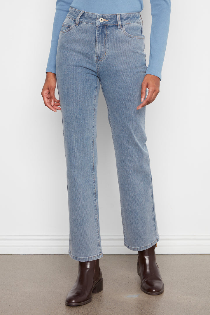 A person wearing light-wash blue straight-leg jeans with a light blue long-sleeve top and dark brown ankle boots, standing against a white wall.