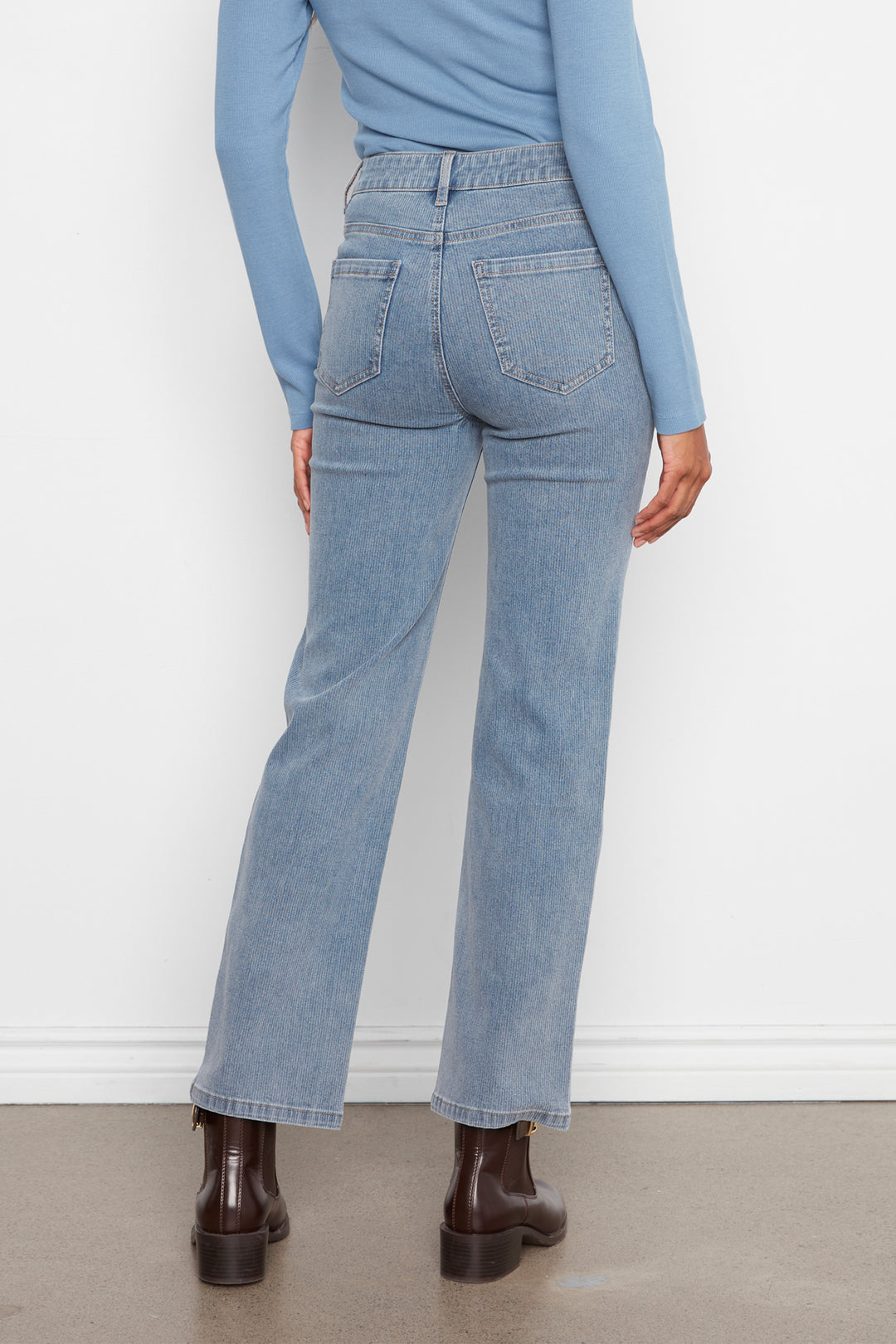 A person shown from the back wearing light-wash blue straight-leg jeans, a light blue long-sleeve top, and dark brown ankle boots against a white wall and gray floor.
