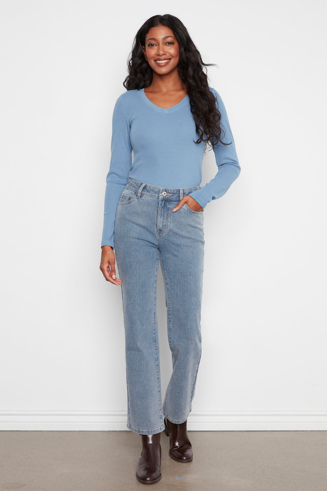 A woman modeling a light blue long-sleeve knit top and light-wash blue straight-leg jeans, standing against a plain white wall.