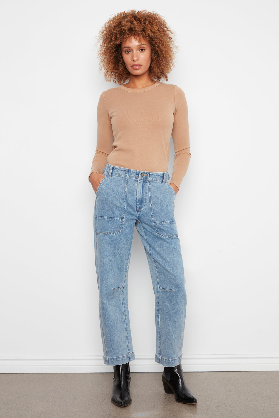 A model with curly light brown hair stands against a white wall wearing a beige long-sleeve crewneck top, light-wash blue high-waisted straight-leg jeans, and black ankle boots.